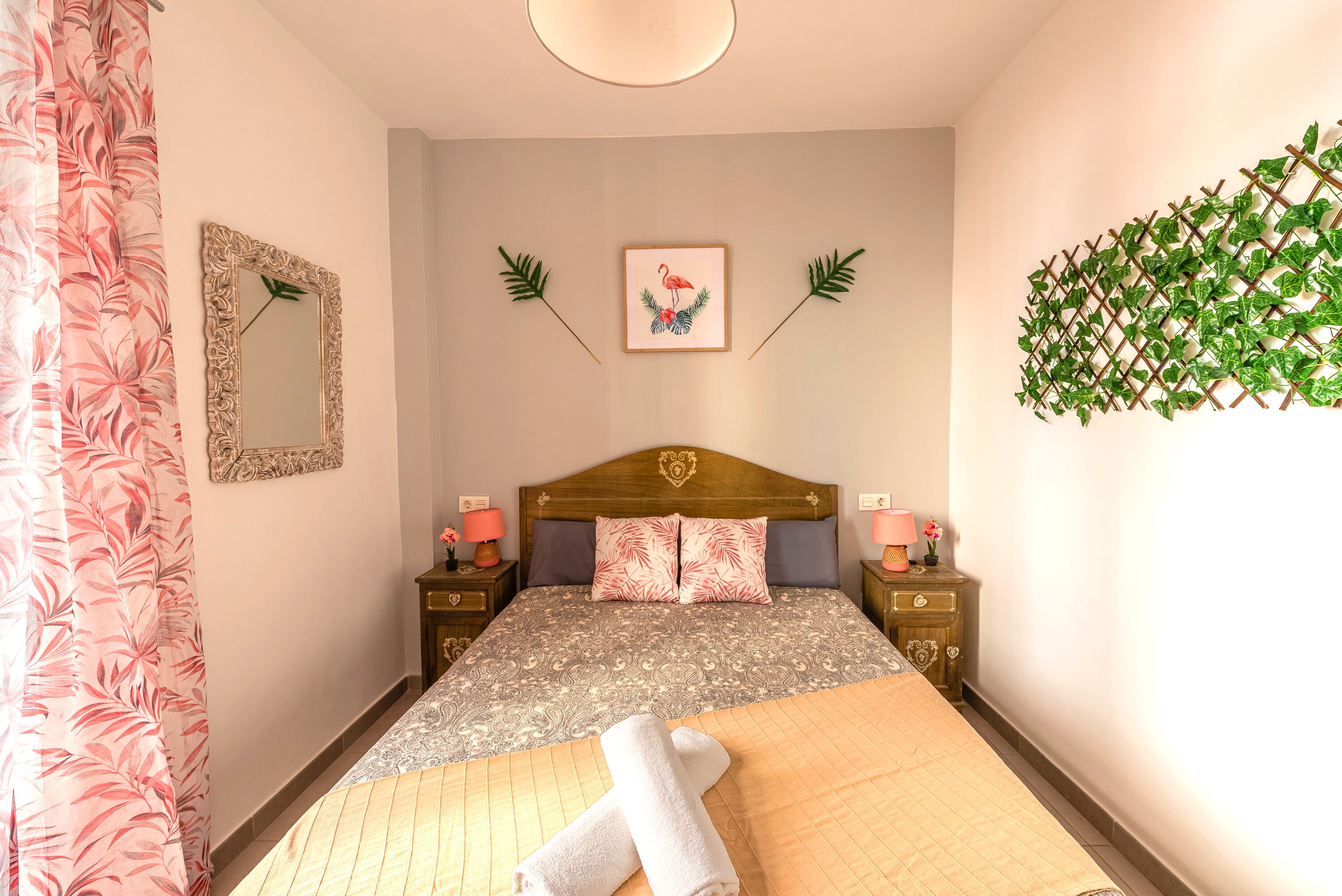 Un dormitorio ordenado con una cama, dos mesitas de noche, un espejo y una planta en la pared.