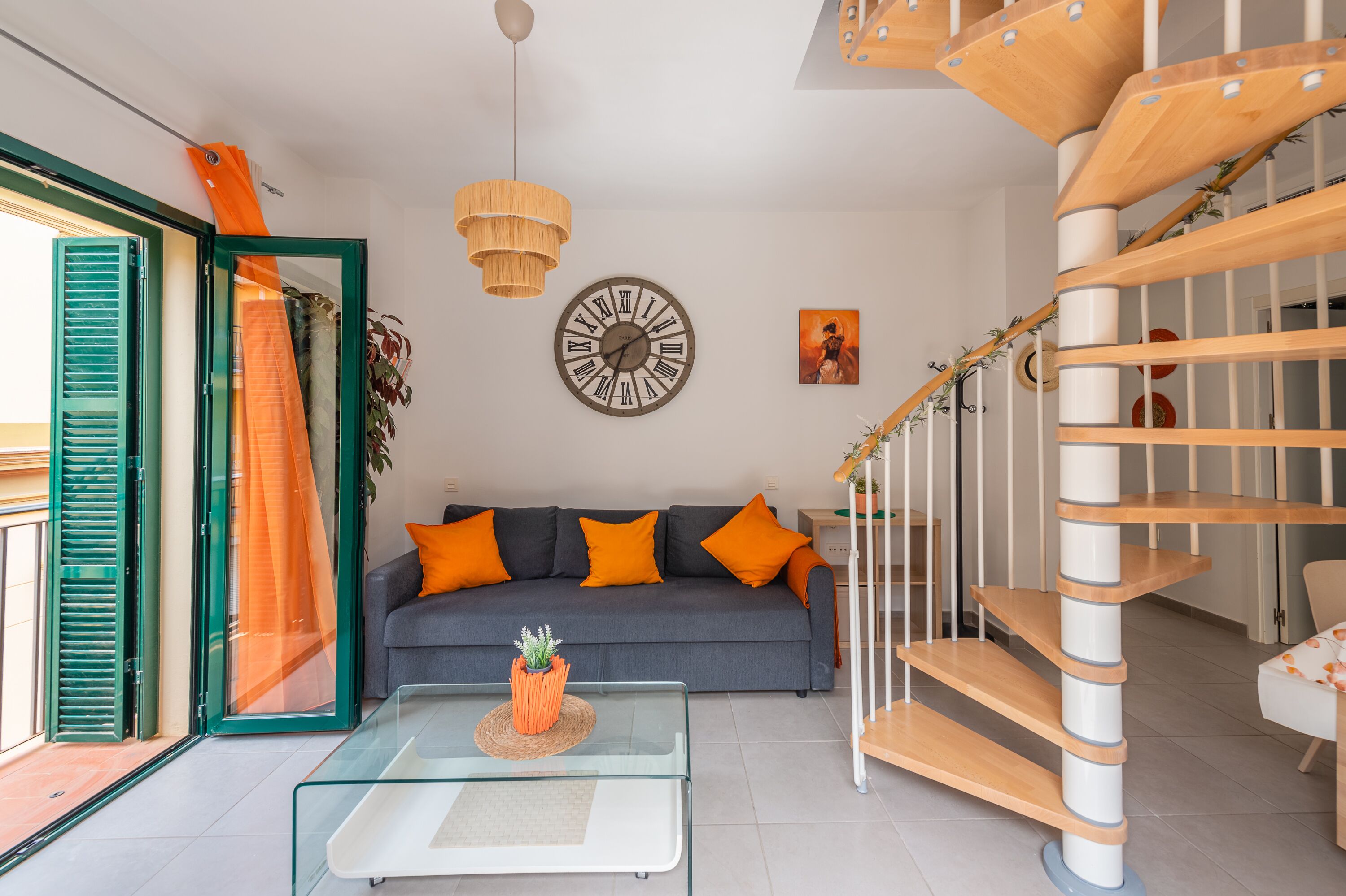 A modern living room with a grey sofa, orange cushions, a wooden spiral staircase, and a round wall clock.