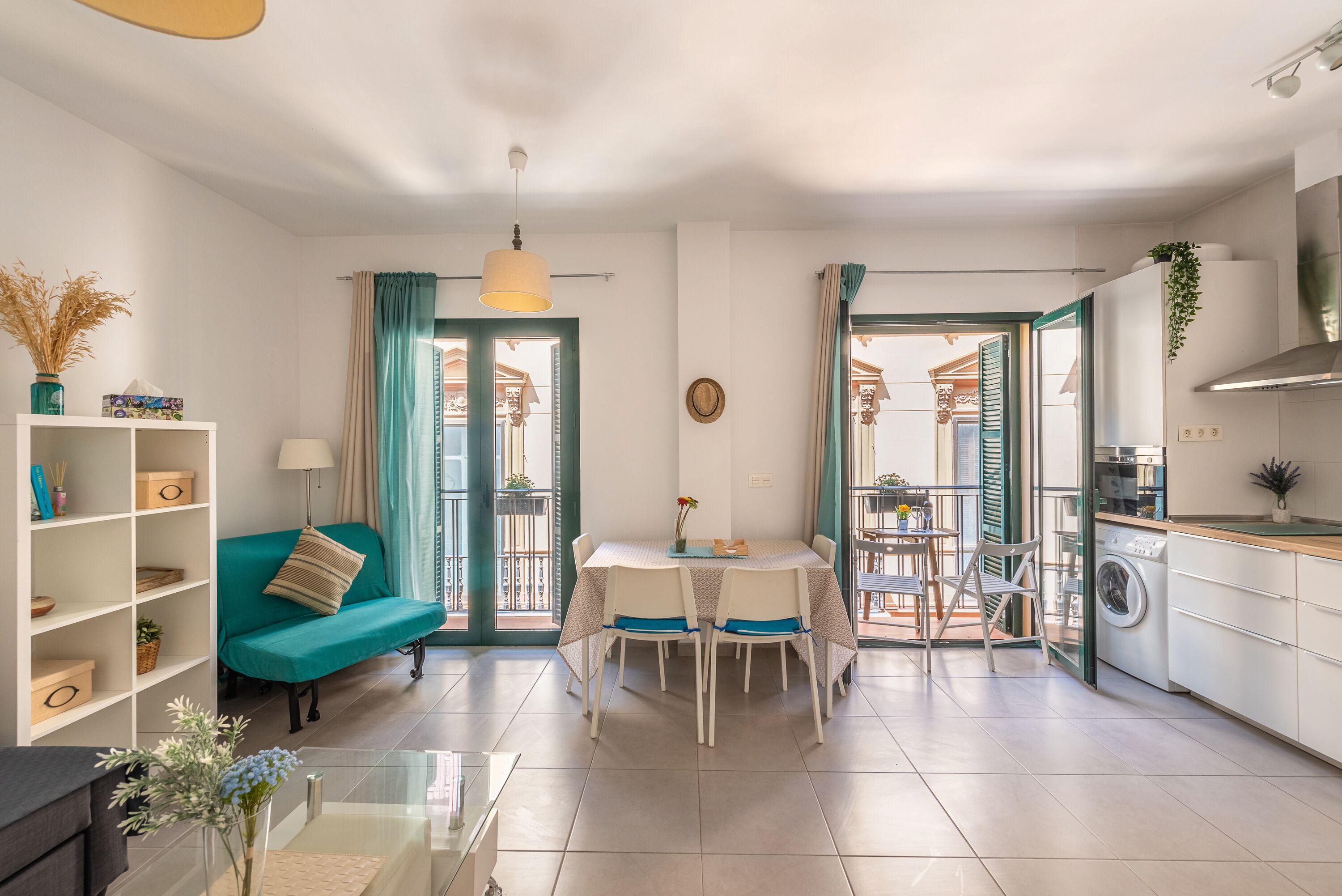 A modern living room with a teal sofa, a dining table with four chairs, a kitchen area with a washing machine, and a balcony with a view.