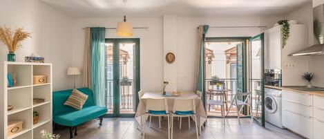 A modern living room with a teal sofa, a dining table with four chairs, a kitchen area with a washing machine, and a balcony with a view.