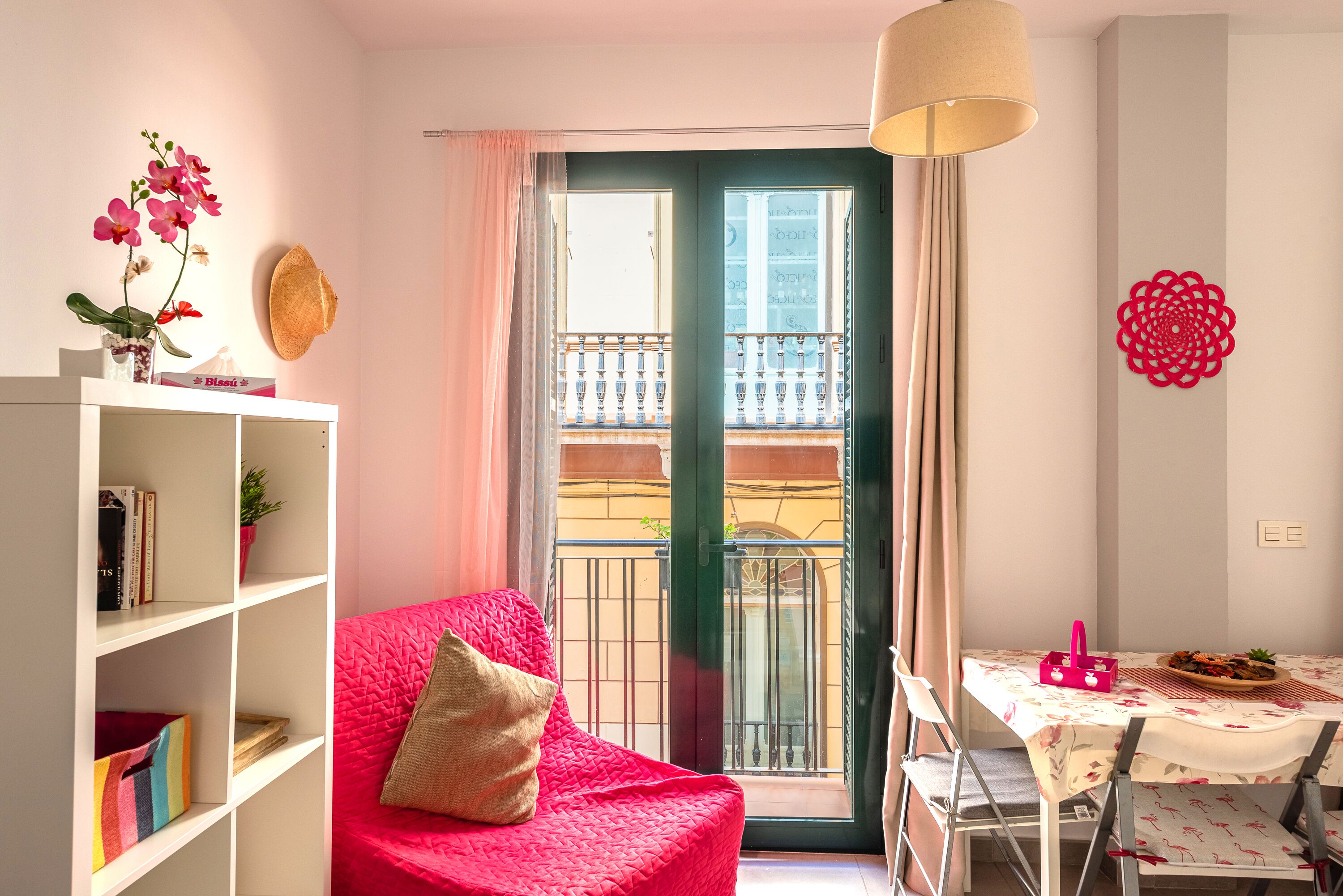 A cozy living room with a red sofa, a bookshelf, a dining table with chairs, a balcony view, and a decorative wall piece.
