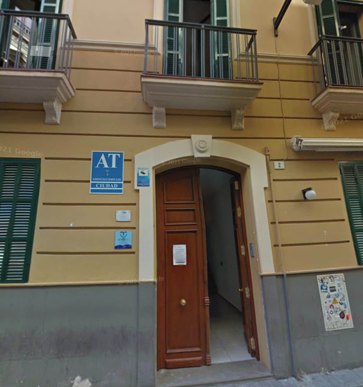 Una entrada a un edificio con una puerta de madera, un letrero azul que dice 'AT' y 'CIUDAD', y una pequeña placa en la pared.