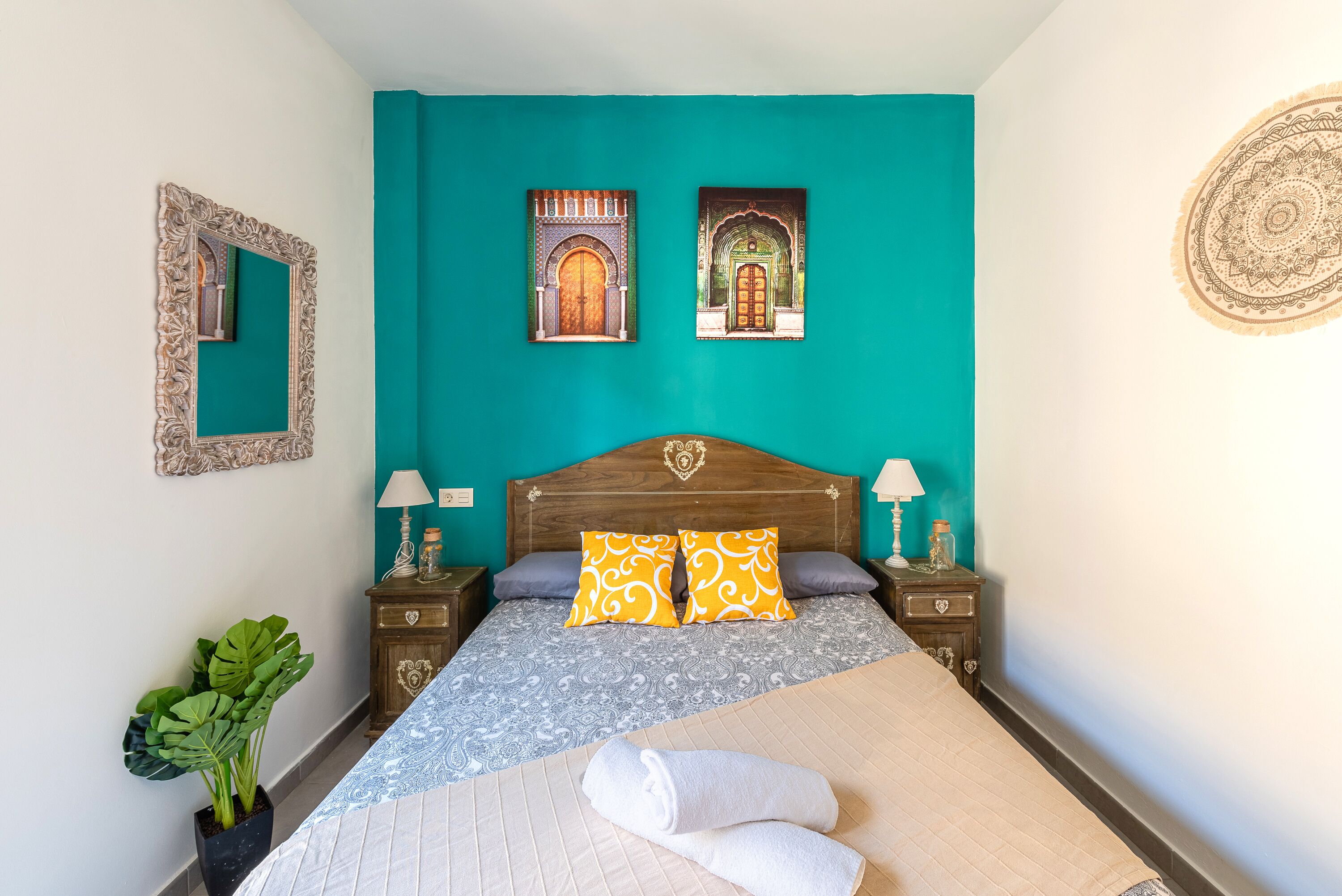 A bedroom with a teal wall, a wooden headboard, two nightstands with lamps, a large bed with patterned bedding, and a potted plant.