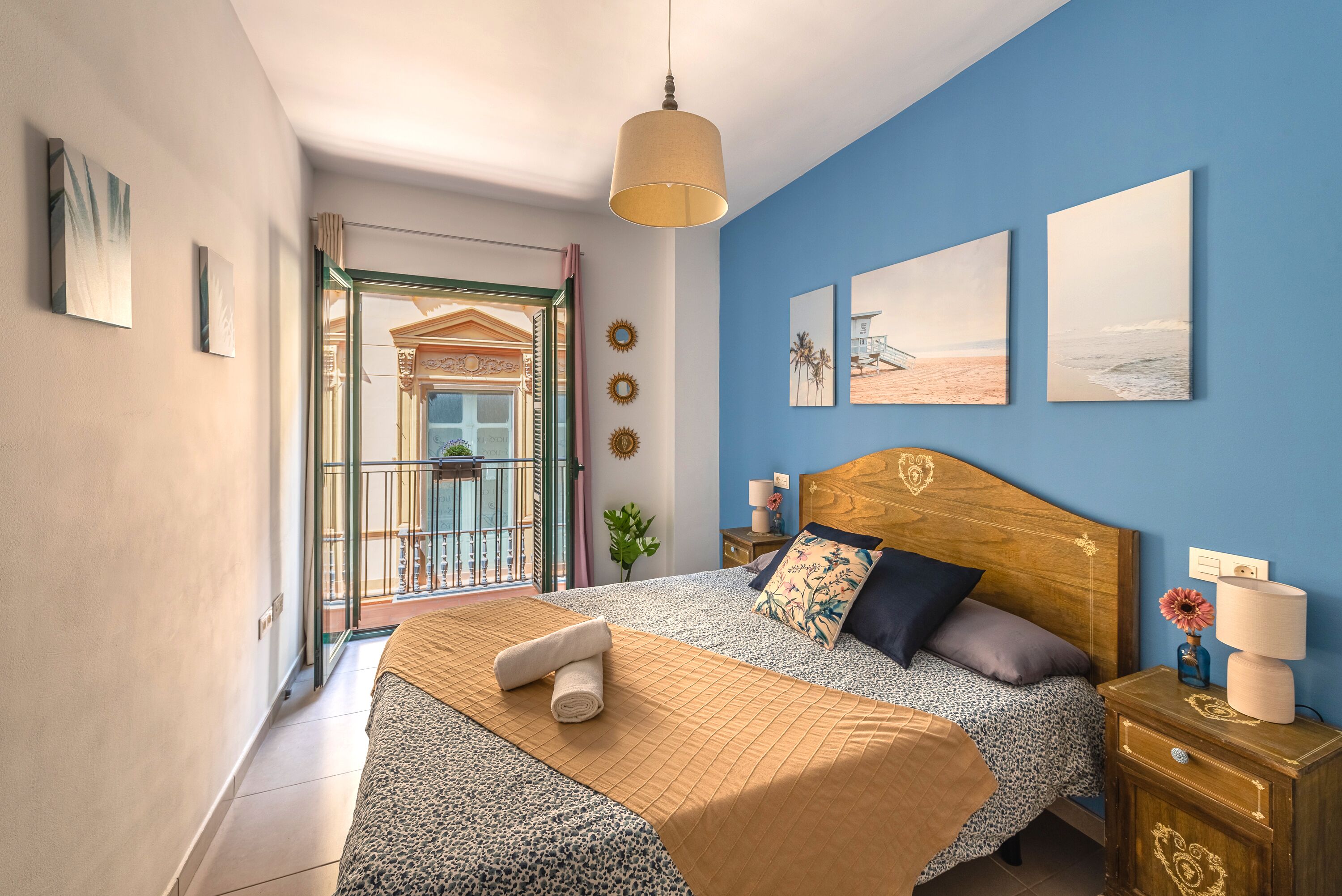Un dormitorio con cabecera de madera, una cama con colcha estampada, una mesita de noche con lámpara y vistas a un balcón.