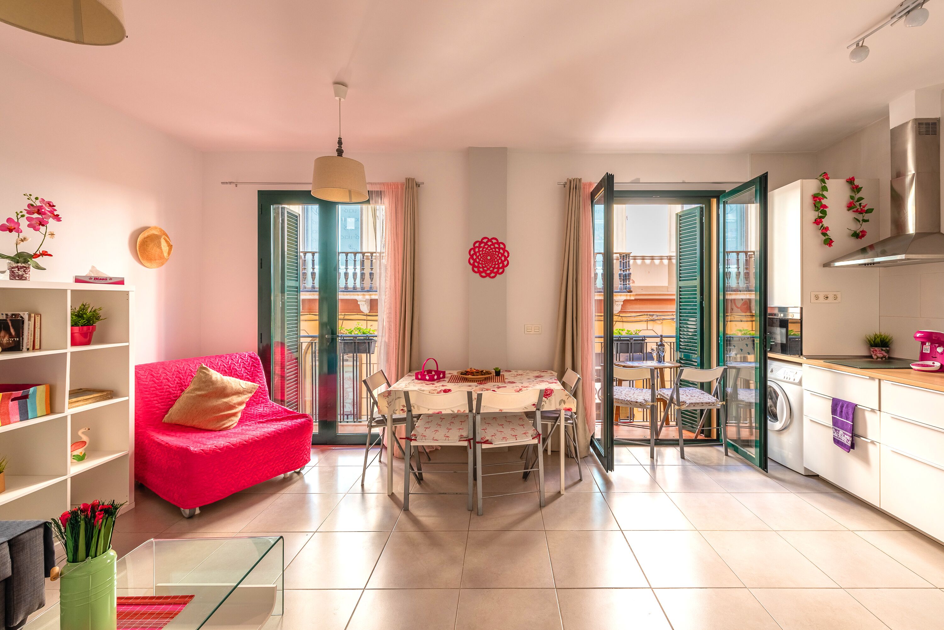 A bright living room with a red sofa, a dining table with chairs, a kitchen area, and a balcony with a view.