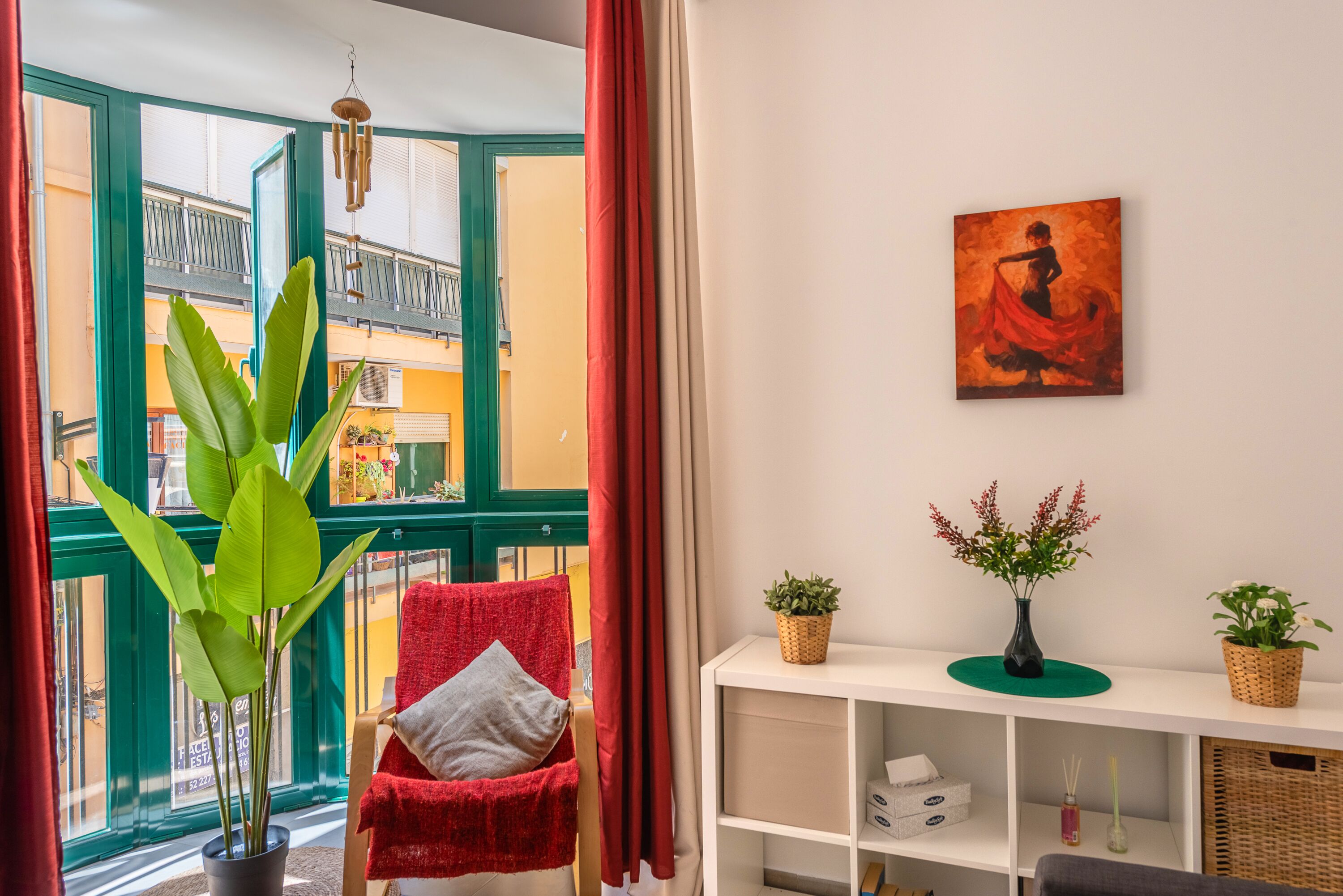 A room with a large window, a red chair, a potted plant, a shelf with decorative items, and a painting on the wall.