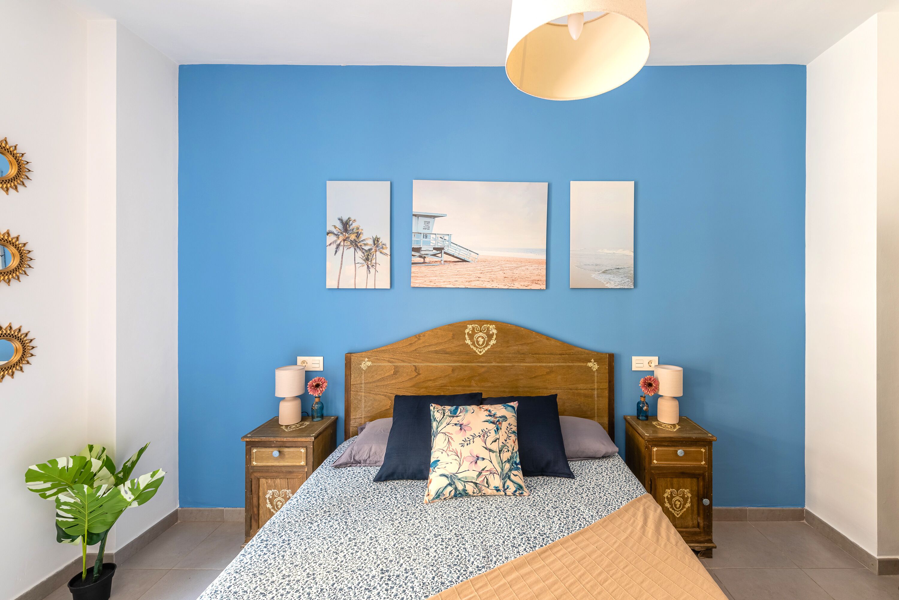Un dormitorio con pared azul, cabecera de madera y cama con ropa de cama estampada. Hay tres cuadros enmarcados en la pared, una planta en maceta y dos mesitas de noche con lámparas.