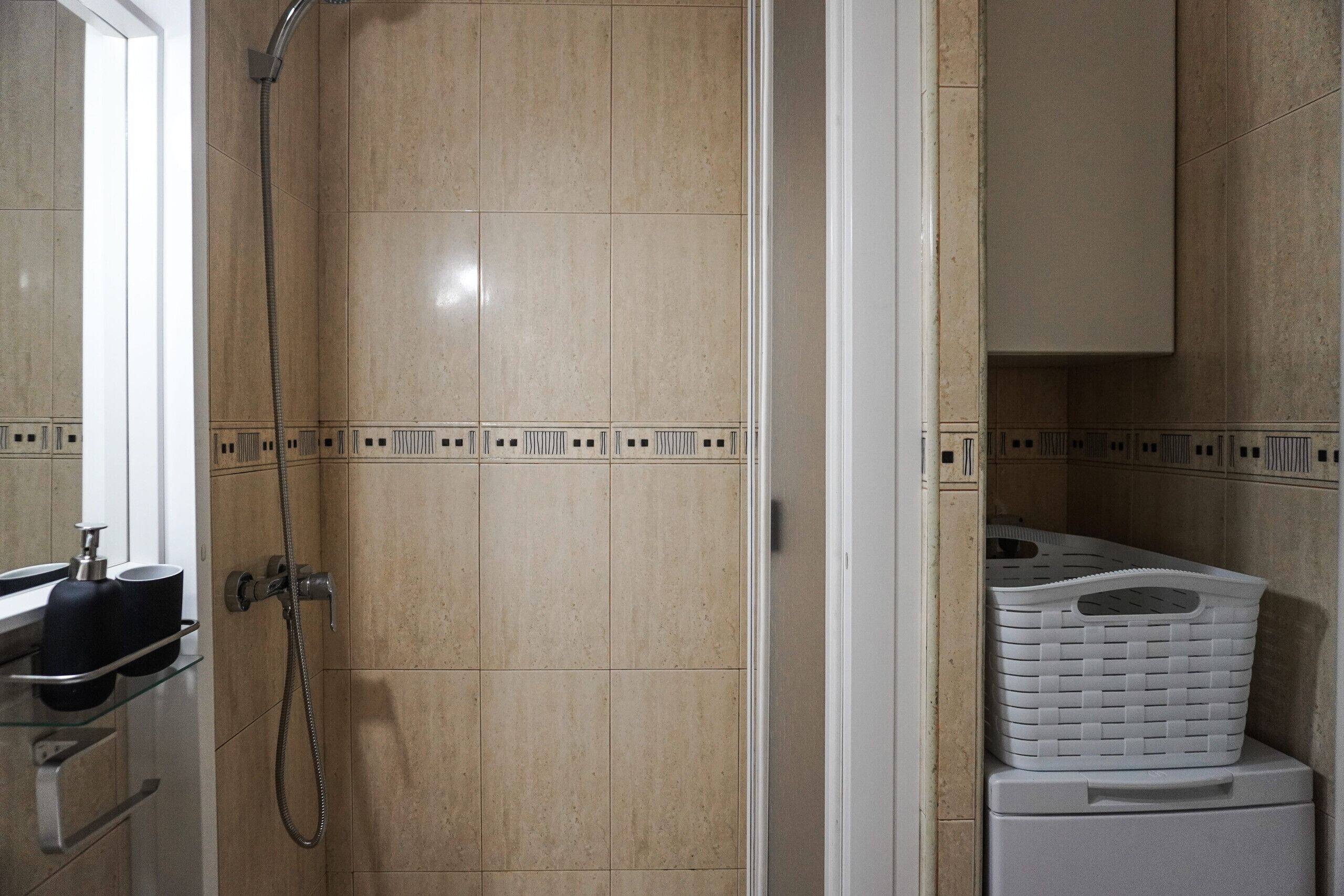 A bathroom with beige tiled walls, a shower with a handheld hose, a white sink with a black soap dispenser, and a white laundry basket on a white cabinet.