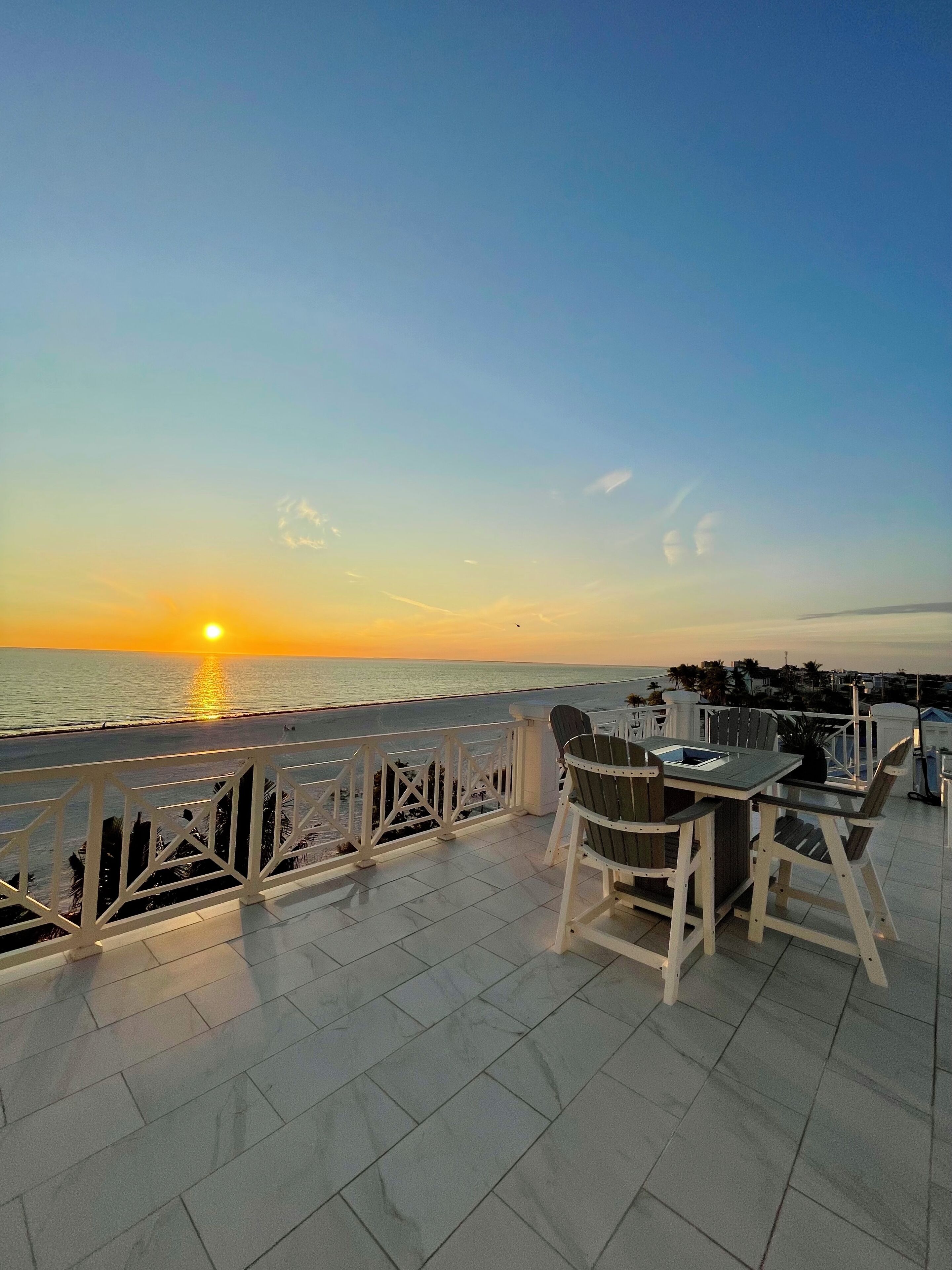 A terrace with a table and chairs overlooking a sunset over the sea.