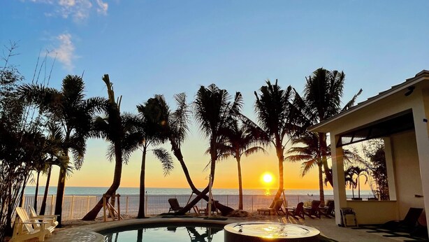 Un espace piscine doté de chaises longues et de palmiers, offrant une vue sur le coucher de soleil sur l’océan.