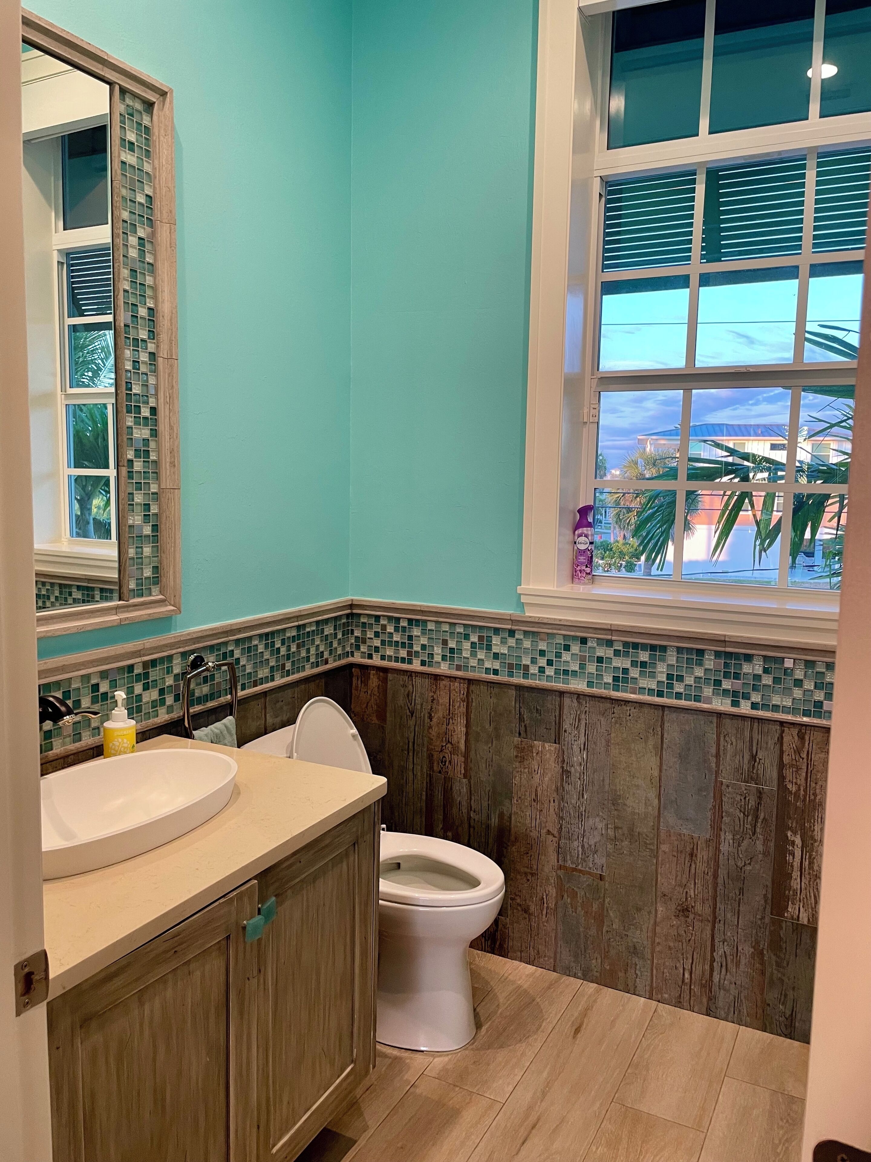 Un baño con pared color turquesa, tocador de madera, inodoro blanco y ventana con vista a palmeras.
