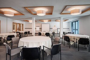 Ein Konferenzraum mit runden Tischen, schwarzen Stühlen und weißen Tischdecken. Deckenleuchten und ein großes Fenster sind im Hintergrund zu sehen.