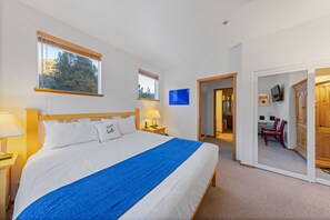 Ein Schlafzimmer mit einem großen Bett, zwei Nachttischlampen, einer blauen Tagesdecke und Blick auf einen Essbereich durch eine offene Tür.