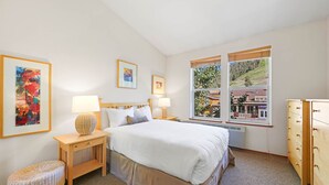 A hotel room with a large bed, two bedside tables with lamps, a small wicker stool, a wooden dresser, and a window with a view of a building and trees.
