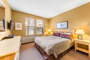 A hotel room with a bed, two bedside tables with lamps, a desk, a television, and two windows with blinds. - 716 | Tahoe 2BD Basecamp - Soaks, Swims + W/D (Olympic Valley)
