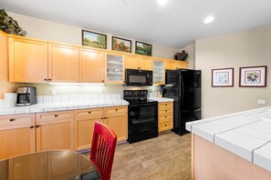 A modern kitchen with wooden cabinets, a black refrigerator, a microwave, and a stove. There are framed pictures on the wall and a red chair in the dining area. - 716 | Tahoe 2BD Basecamp - Soaks, Swims + W/D (Olympic Valley)