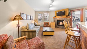 A living room with a sofa, armchairs, a coffee table, a fireplace, a TV mounted above it, and a dining area with a table and chairs.