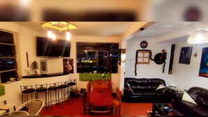 Un salon avec un canapé en cuir noir, un fauteuil rouge, une table à manger avec des tabourets de bar et une grande fenêtre donnant sur la ville.