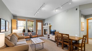 A spacious living room with a fireplace, a dining area with wooden furniture, and large windows offering a view of the outdoors. - 712 | Outdoor Lover's 2BD - King, Bunks + Pool (Olympic Valley)