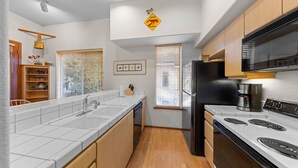 A modern kitchen with white cabinetry, a black refrigerator, a stove, and a microwave. There is a wooden shelf with decorative items and a window with blinds. - 521 | Roomy 2BD/2BA - Kitchen, W/D, Pool + Spas (Olympic Valley)