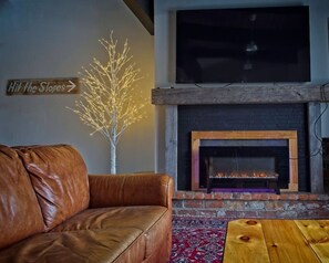 A living room with a leather sofa, a fireplace, a flat-screen TV, and a decorative sign.