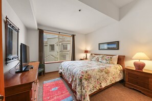 Une chambre à coucher avec un lit, un bureau avec un ordinateur, une fenêtre avec des stores, une table de chevet avec une lampe et un tableau encadré au mur.