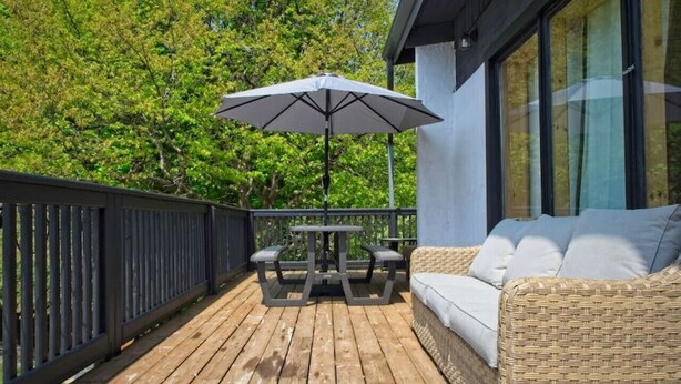 A wooden deck with a table and chairs under a large umbrella, a wicker sofa with cushions, and a building with large windows in the background.