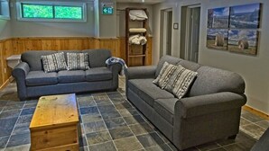 A room with two grey sofas, a wooden coffee table, and a stone-tiled floor. There is a window with a view of greenery, a shelf with towels, and framed pictures on the wall.