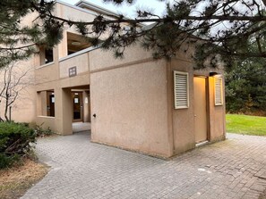 Un bâtiment moderne avec un chemin en briques menant à un ensemble de portes, le tout entouré d’arbres.
