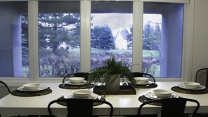 A dining table set with white plates and bowls, centered under a pendant light fixture, with a view of a house and trees through large windows.