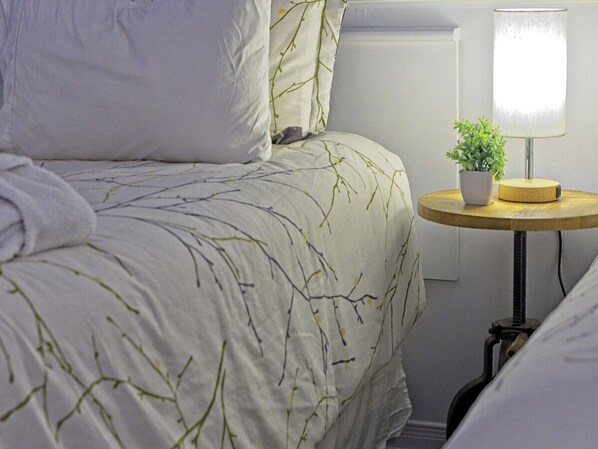 Una cama bien hecha con estampado de ramas de árbol, una mesita redonda con una planta y una lámpara.