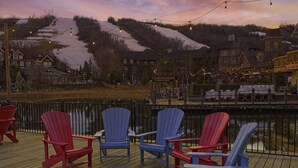 A deck with Adirondack chairs, a wooden floor, and a view of buildings and a snowy landscape.
