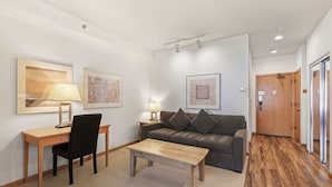 Una sala de estar moderna con un escritorio de madera, una silla negra, un sofá gris y una mesa de centro de madera. Hay dos obras de arte enmarcadas en las paredes y una puerta de madera a la derecha.