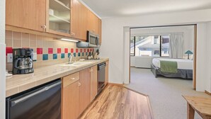 Cocina compacta con armarios de madera, microondas y cafetera. Se observa un pequeño comedor con mesa y una cama al fondo.