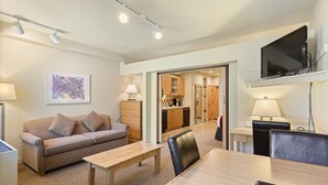 A modern living room with a sofa, coffee table, and dining table. There is a kitchen in the background with cabinets and a television mounted on the wall. - 250 | Easy-Breezy Studio -AC, Spas, Pool & Tennis (Olympic Valley)