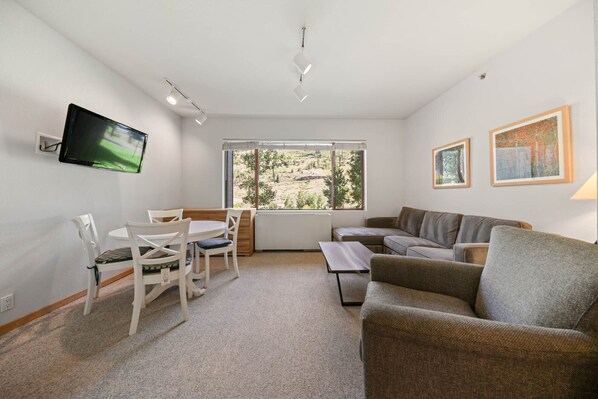A living room with a sofa, a dining table with chairs, a TV mounted on the wall, and a window with a view of trees.