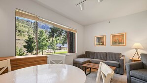 A modern living room with a round table, a sofa, and a couch. There is a large window with a view of a mountainous landscape.