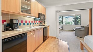 A modern kitchen with wooden cabinets, a coffee maker, and a dishwasher. There is a dining area with a table and chairs, and a view of the outdoors through a large window.