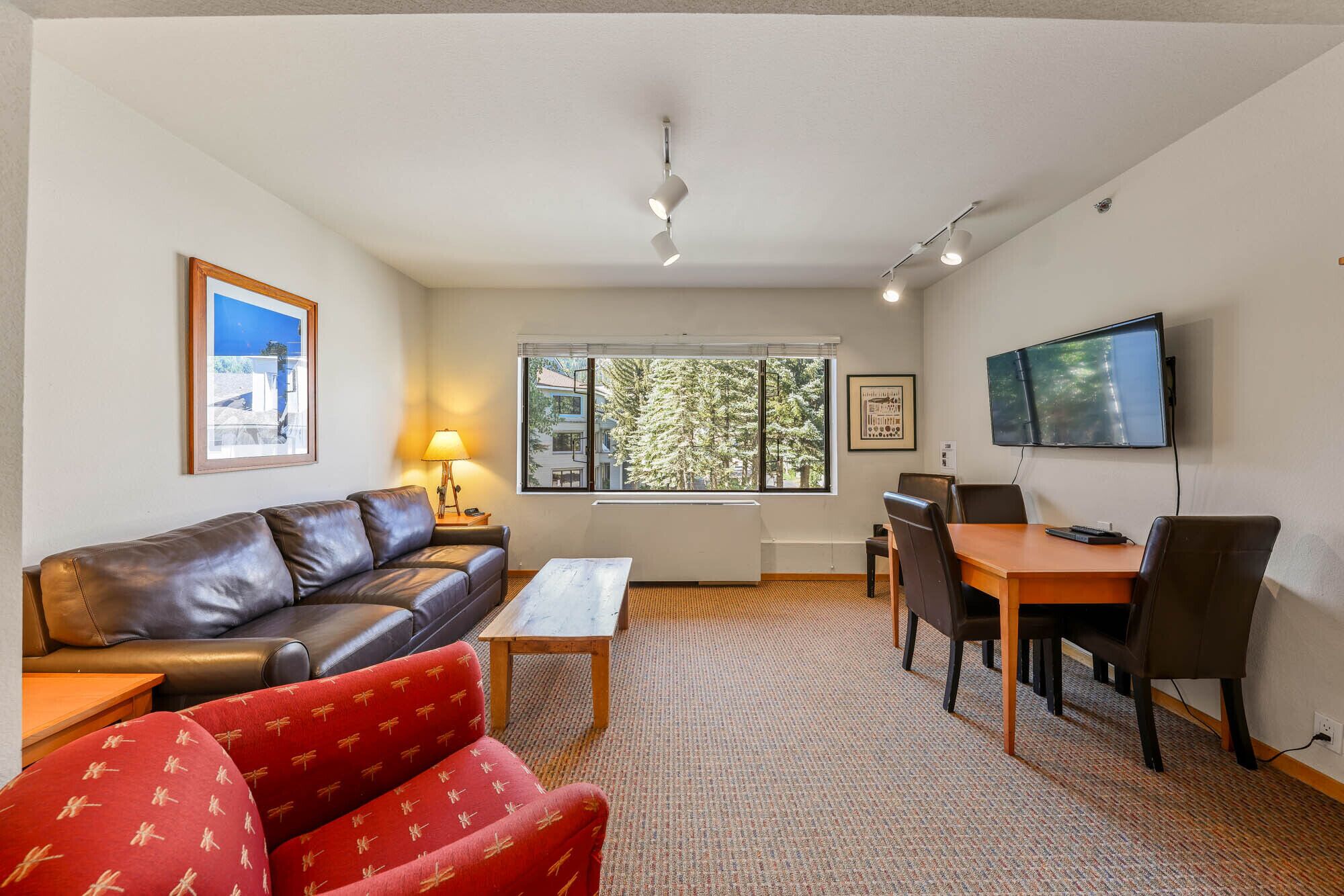 A living room with a leather sofa, a red patterned chair, a wooden coffee table, a dining table with chairs, a TV mounted on the wall, and a window with a view of trees.