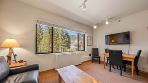A modern living room with a sofa, a wooden coffee table, a dining table with chairs, a TV mounted on the wall, and a large window with a view of trees and buildings. - 246 | Scenic Studio - Hit Trails, Then Hot Tubs (Olympic Valley)