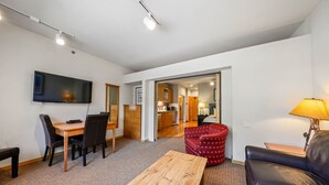 A modern living room with a flat-screen TV, a wooden coffee table, a red patterned armchair, a black leather sofa, and a kitchen area visible through an open door. - 246 | Scenic Studio - Hit Trails, Then Hot Tubs (Olympic Valley)
