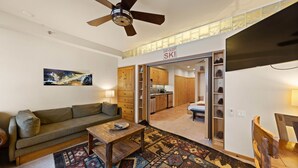 A cozy living room with a sofa, coffee table, and a rug. A kitchen is visible through an open door, and there's a ceiling fan. - 243 | Premium Studio - Mtn Views + Pool Escape (Olympic Valley)