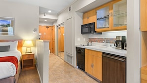 Una habitación de hotel compacta con una cama, mesita de noche, lámpara y una kitchenette equipada con microondas, cafetera y armarios.