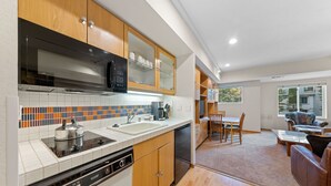 A modern kitchen with wooden cabinets, a dishwasher, and a microwave. There is a dining area with a table and chairs, and a sofa in the living room. - 232 | Updated Studio w/AC! - Pool View + Parking (Olympic Valley)
