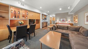 A modern hotel room with a kitchenette, a dining area, a living room with a sofa and a coffee table, and a bedroom with a bed and bedside lamps.
