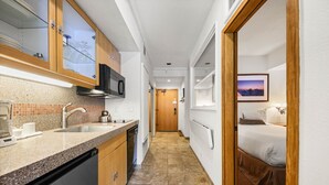A narrow hallway with a kitchen area on one side, a bed on the other, and a door leading to another room.