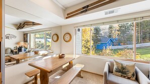 A cozy room with a wooden dining table, a sofa with a decorative pillow, and a large window offering a view of a blue house and trees.