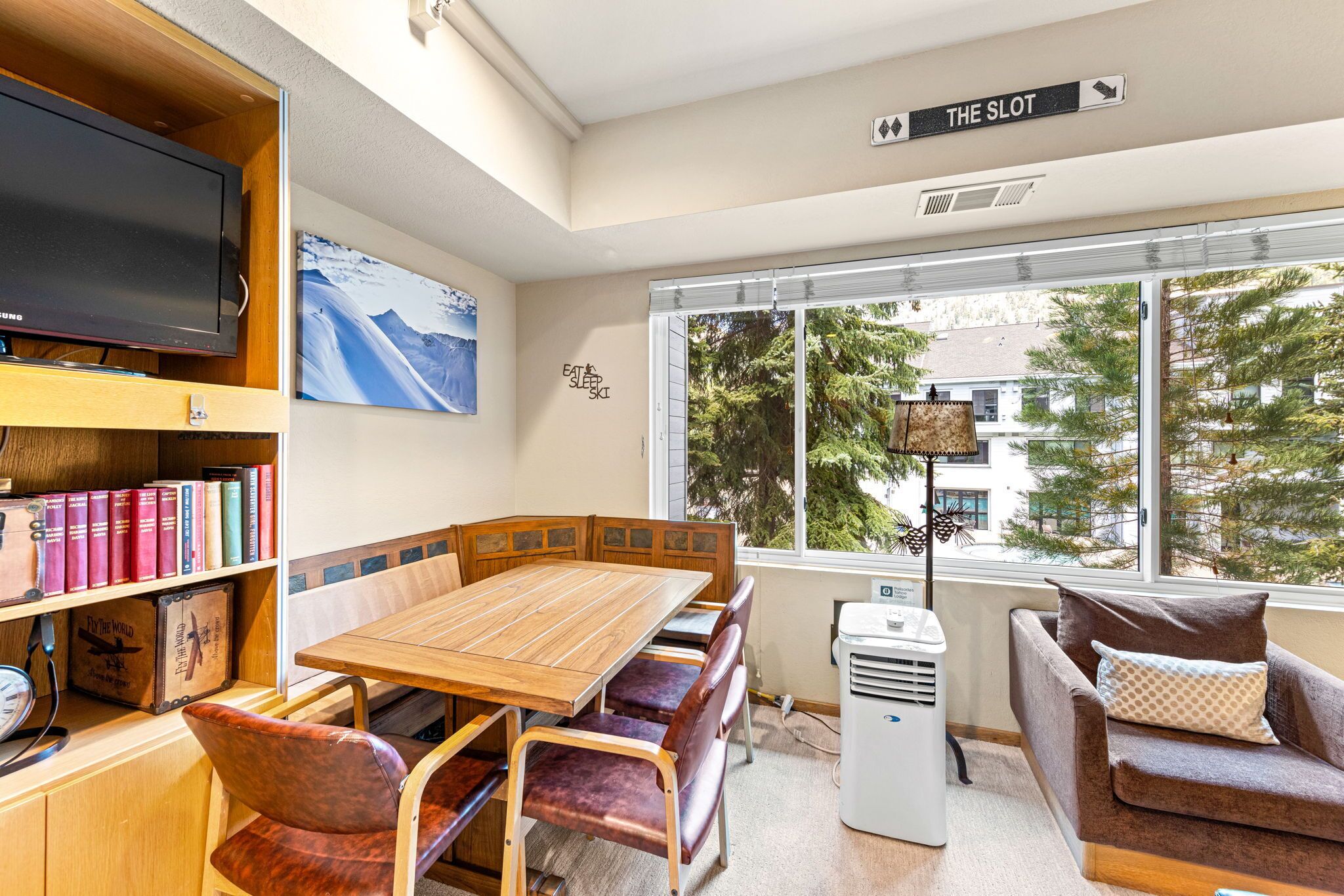 A cozy room with a wooden table, chairs, a bookshelf, a television, and a large window with a view of trees and buildings.