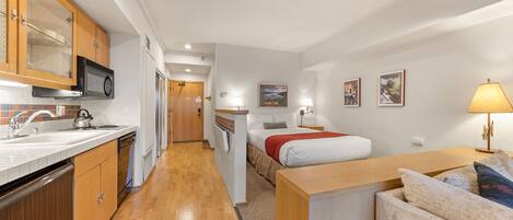A compact hotel room with a kitchenette, a bed with a red blanket, a small table, and a sofa.