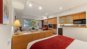 A modern bedroom with a bed, a wooden dresser, a desk, and a kitchenette.